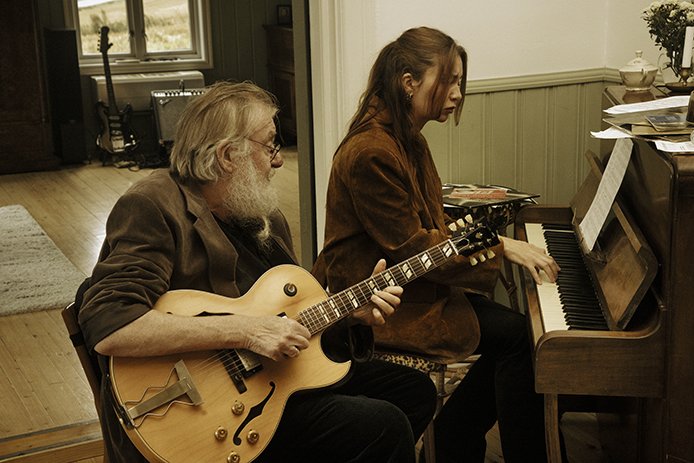 En eldre mann med langt skjegg sitter og spiller gitar mens han ser på en yngre kvinne som spiller piano